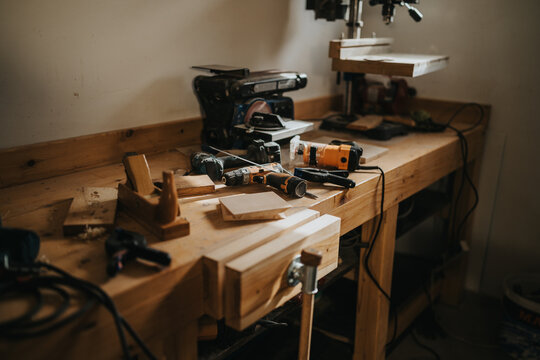 Woodworking workbench filled with power tools, hand tools and wood pieces. A carpentry workshop scene showing tools, drill press and unfinished lumber on a bench.