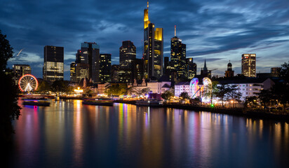 Obraz premium Frankfurt skyline illuminated at dusk with colorful reflections on Main river, modern skyscrapers and ferris wheel in urban waterfront scene
