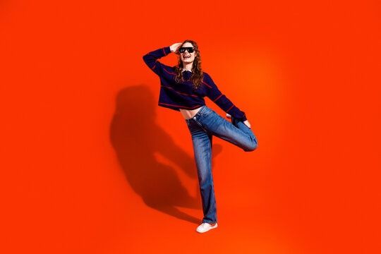 Happy young woman in sunglasses and jeans striking a playful pose against a bright orange studio background