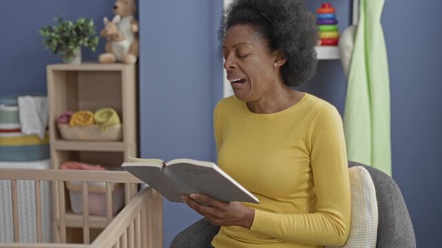 Woman holds book and reads to cradle with hands by a crib in a building nursery, wearing yellow sweater and seated beside the crib; comfort bonding.