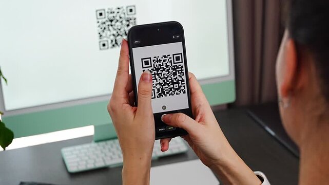 Person scanning QR code from computer screen with smartphone at home workspace. Digital interaction between devices and online service access concept.