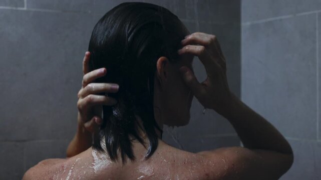 Slow motion static shot of person washing hair and rinsing shampoo in dim tiled shower from behind