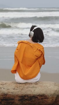Static shot of woman adjusting orange jacket while sitting on driftwood at ocean beach with headphones during overcast day