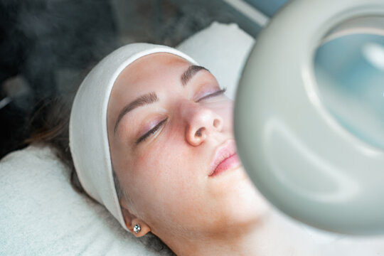 A young woman with eyes closed wearing a headband lies down during a professional facial treatment involving steam and a magnifying lamp at a beauty clinic.