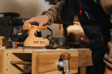 Carpenter uses an electric orbital sander to smooth a wooden board on a workbench. The close-up scene shows hand-held sanding in a warm, tool-filled workshop environment. © qunica.com