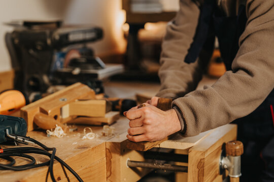 A carpenter uses a hand plane to smooth a wooden board at a workbench. The close-up shows hands, wood shavings and tools in a warm workshop setting.