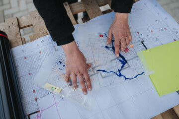 Hands of a planner point to a river and features on architectural blueprints and layered site maps. A professional reviews overlapping maps and technical drawings during a planning session. © qunica.com