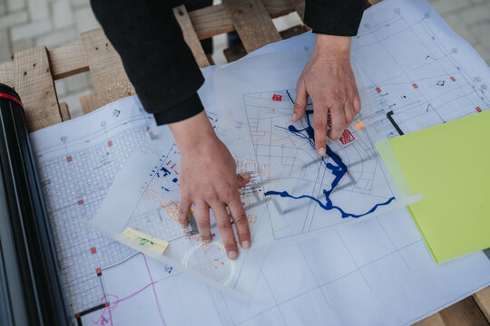 Hands of a planner point to a river and features on architectural blueprints and layered site maps. A professional reviews overlapping maps and technical drawings during a planning session.