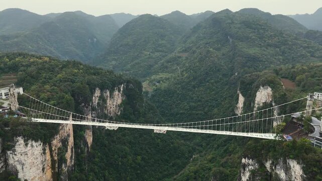 Crossing glass suspension bridge in Zhangjiajie, China. Transparent walkway crosses lush mountain canyon. Suspended glass bridge connecting cliffs over misty valley