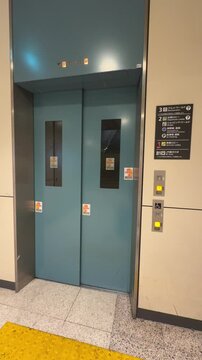 blue elevator doors inside a public building, with narrow vertical windows and multiple stickers attached. control buttons and a floor directory sign in Japanese are mounted on the wall beside it