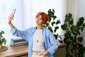 Young woman with vibrant red hair takes a cheerful selfie in a cozy home setting