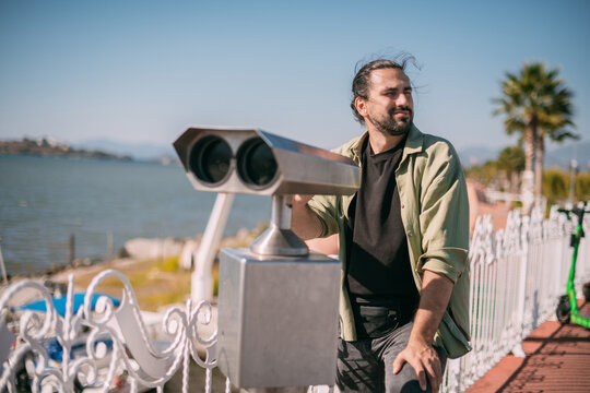A young male tourist looks through large street binoculars on the coast by the sea.