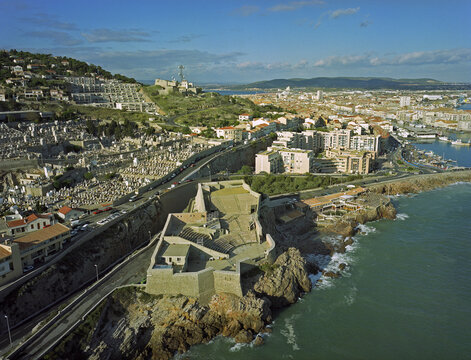 Aerial view of the rugged coastline meets the historic Theatre de la Mer, juxtaposed against the sprawling city and verdant hills under a bright sky, Marseille, France.