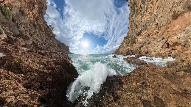 A panoramic wide-angle view of a rocky coastal scene, where waves surge against jagged formations under a bright sun and textured cloud patterns in the sky