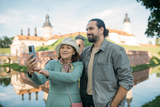 A pensioner and her adult son on a guided tour of an ancient castle. They smile happily and take selfies on their phones. An elderly mother and son travel together, visiting historical sites