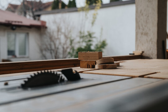 A table saw blade and several wooden offcuts rest on a workbench in a workshop. The close-up composition shows woodworking tools and cut lumber on a bench surface.