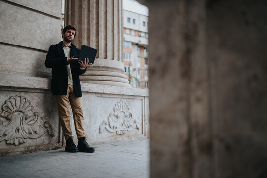 A businessman stands by a stone column holding a laptop and looking thoughtful. A man works outdoors in an urban architectural setting with his laptop.