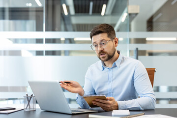Fototapeta premium Professional man wearing glasses engaged in an online video conference, discussing business strategies while holding a notebook and pen, working from a modern corporate office