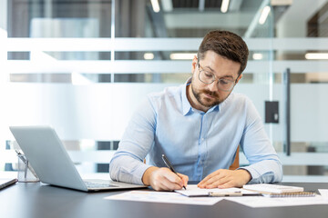 Naklejka premium Businessman wearing glasses and a light blue shirt concentrating on writing notes on a document while working at a desk, with a laptop and various papers in a modern office environment