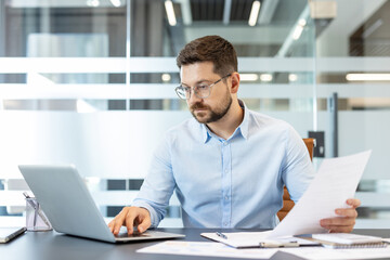Fototapeta premium Businessman wearing eyeglasses and a blue shirt, concentrating on his laptop and carefully reviewing paperwork while sitting at a desk in a modern corporate office environment