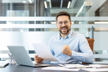 Obraz premium Happy businessman cheering with a fist pump and a wide smile, holding official document in hand while sitting at a desk with a laptop and paperwork, celebrating good news and career achievement
