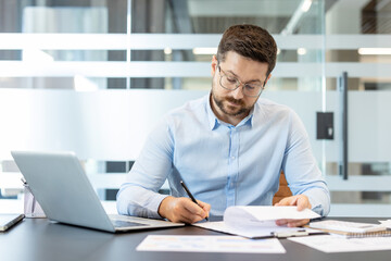 Fototapeta premium Businessman in glasses sitting at office desk, reviewing and signing paperwork, working on a laptop with focus and concentration, managing corporate tasks