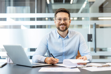 Fototapeta premium Young man in glasses and blue shirt doing paperwork at his desk, holding a pen and looking at camera with a cheerful expression in a modern corporate workspace setting