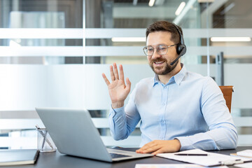 Fototapeta premium Man wearing a headset and spectacles, smiling and waving hand to laptop screen, participating in a remote work virtual meeting from his modern office desk