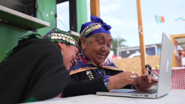 Young indigenous woman teaching an elder how to use a laptop at a table outside a food truck
