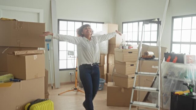 Woman with arms outstretched amid packed moving boxes, ladder and yellow suitcase in new home building; fresh start joy.
