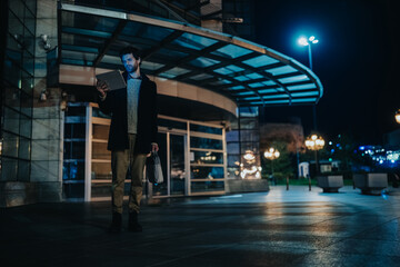 Fototapeta premium A businessman stands outside an office building at night looking at a tablet. He checks his tablet under a modern canopy in a quiet urban plaza.