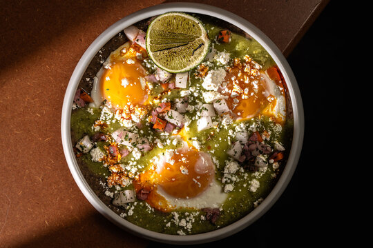 Overhead view of a Mexican style egg dish with salsa verde, crumbled cheese, diced vegetables and grilled lime. Strong directional light creates contrast and highlights textures.
