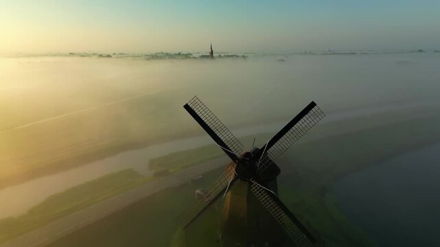 Aerial view of a windmill surrounded by fog and a distant village with a church tower, creating a serene atmosphere, Schermerhorn, North Holland, Netherlands.