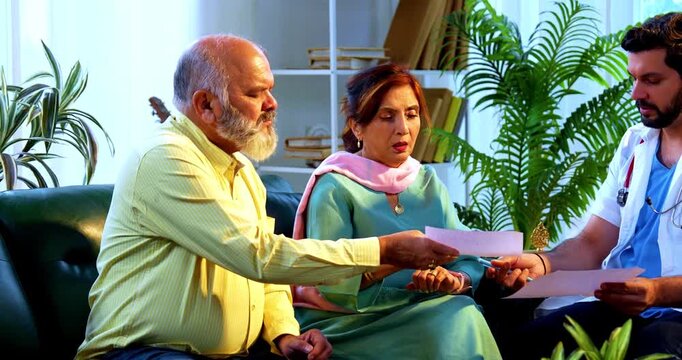 Professional Indian young male doctor explaining medical case to Asian senior retired couple patients sitting on a sofa in a modern clinic; healthcare, medical advice, and old age care concept.