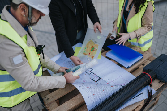 Construction engineers in safety vests examine blueprints and site plans on a wooden pallet. The team reviews architectural drawings and project documents during an on-site planning meeting.