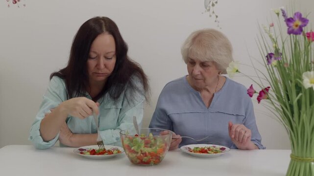 Two women eating calmly at dining table. Pair of women quietly dining with salad between them. Two Caucasian women share tranquil moment while eating together at bright dining space