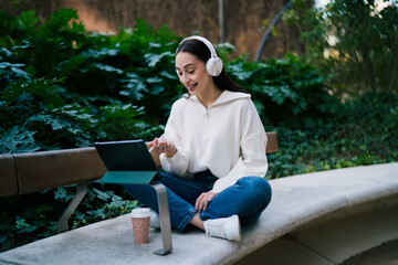 Fototapeta premium Young smiling woman in headphones sitting on park bench and looking at digital tablet. Happy student girl using mobile device for online learning or entertainment in green urban environment.