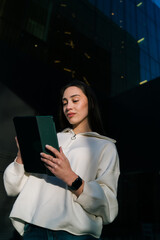 Serious woman with braces checking digital content on tablet outdoors against dark office background. Female user browsing internet or reading news on mobile device, connectivity and modern tech. © BullRun