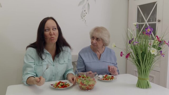 Women share salad. Caucasian women converse over salad. Women at dining nook savoring and discussing salad. Two women seated at table taste and chat about their fresh salad meal