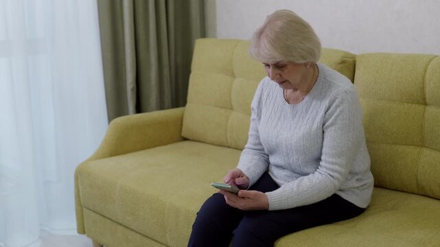 Elderly woman connecting. Older lady engaging with smartphone in cozy room. Senior Caucasian woman sitting comfortably in welllit living area while discovering new texting app
