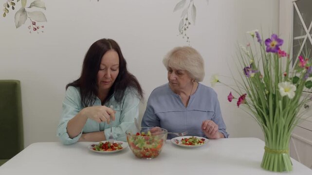 Two Caucasian females relaxing with food and gentle talk inside home. Two women of European descent engaged in peaceful meal sharing amidst cozy floral setting and friendly exchange