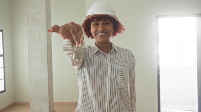 Woman holding keys with outstretched hand in an empty building room, smiling while wearing hardhat and striped shirt as architect presenting access; homeowner pride.