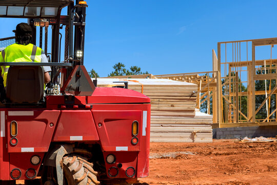 Forklift operator moves stacks of lumber on construction site where workers build new structures outside.