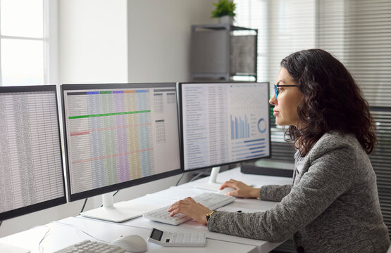 Analyst woman performing data analysis at office computer. She reviews spreadsheet on triple monitor screens, managing finance reporting, verifying KPI. Business analytics and reporting concept.