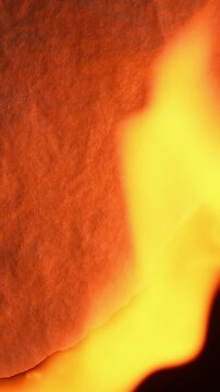 Burning paper with flames spreading across textured surface, close up fire consuming sheet on dark background, abstract dramatic scene with copy space for vertical video format