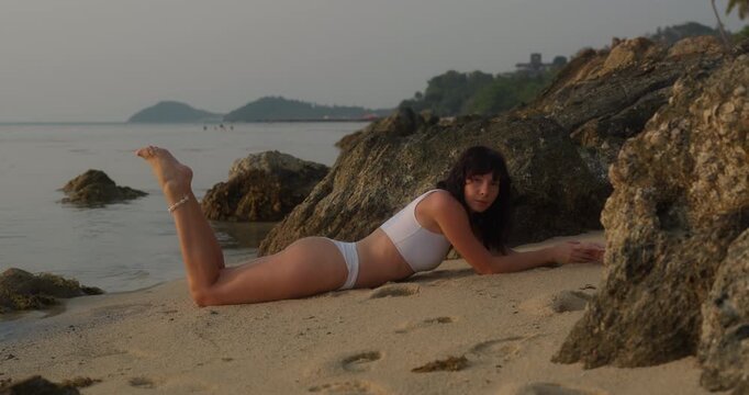 Portrait of carefree barefoot woman lies on stomach and swings legs on sand beach by rocks. Pretty female in white bikini rests on ocean coast at exotic resort