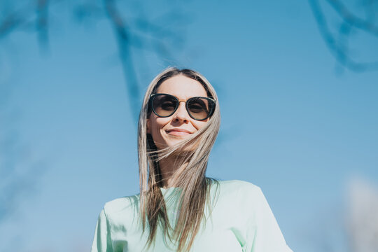 Young woman wearing sunglasses smiles at the camera against a clear blue sky. Casual portrait shows her enjoying sunny outdoor weather with hair blowing in the breeze.