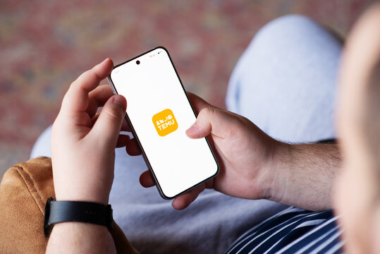 Kyiv, Ukraine. April 3, 2026. Man holding a smartphone displaying the Temu app icon on a white screen while sitting indoors.