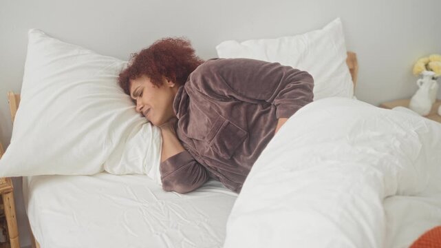 Woman clutching stomach on bed in building, hands pressing abdomen under white duvet and orange blanket; discomfort rest.