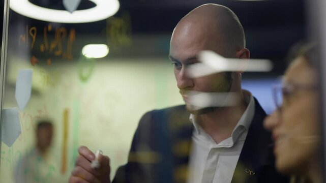 A focused bald businessman writes ideas on a transparent glass whiteboard during a collaborative session. He outlines plans and strategies with a marker in a modern office environment.